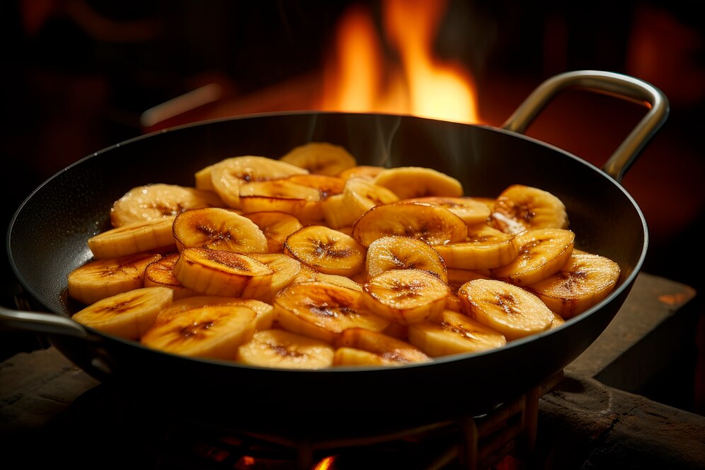Sizzling sliced bananas in a black frying pan, with steam rising against a warm, glowing fire backdrop.