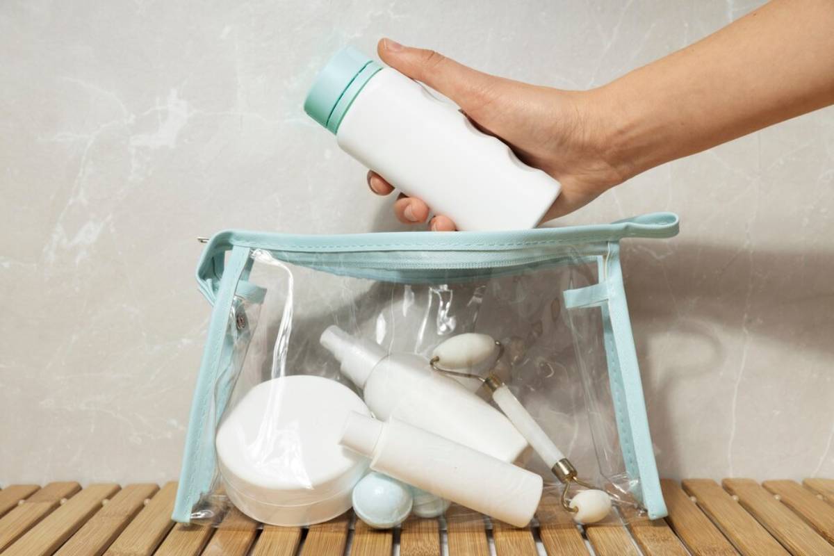 A hand places a white bottle into a clear cosmetic bag filled with various hand sanitiser products on a wooden surface.