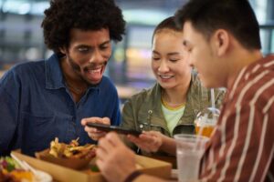 Three friends gather around a table with food, sharing a phone, and enjoying drinks in a vibrant, casual dining setting.