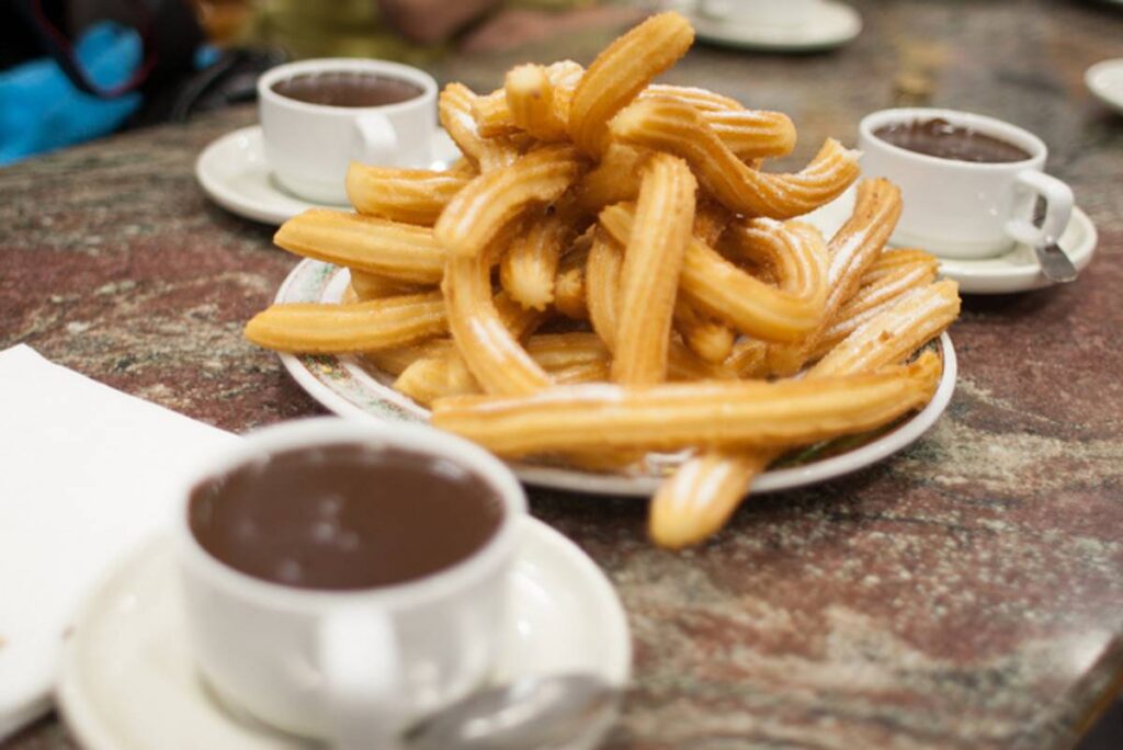 A plate of golden, crispy churros piled high, accompanied by small cups of rich dark chocolate for dipping.