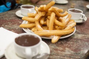A plate of golden, crispy churros piled high, accompanied by small cups of rich dark chocolate for dipping.