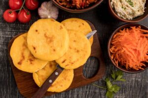 A wooden board holds several golden arepas near fresh tomatoes, garlic, shredded carrots, and bowls of seasoned meat and salad.