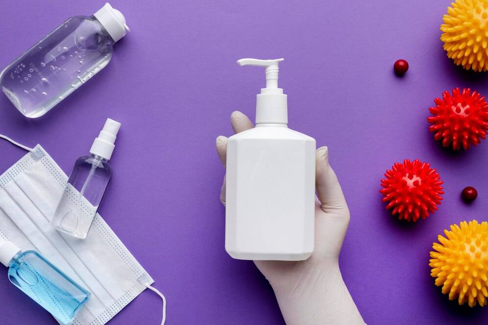 A gloved hand holds a white pump bottle among sanitizers, a mask, and colorful virus models on a purple background.