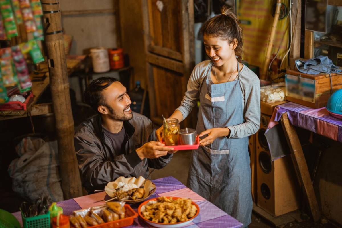 A woman in an apron serves drinks on a tray to a seated man in a casual eatery filled with colorful food offerings.