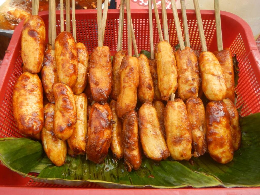 A basket filled with skewered, caramelized banana fritters on banana leaves, showcasing their golden, crispy texture.