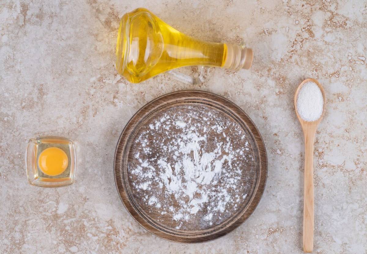 A wooden plate with flour marks, a cracked egg in a glass, a spoon of salt, and a bottle of oil on a textured surface.