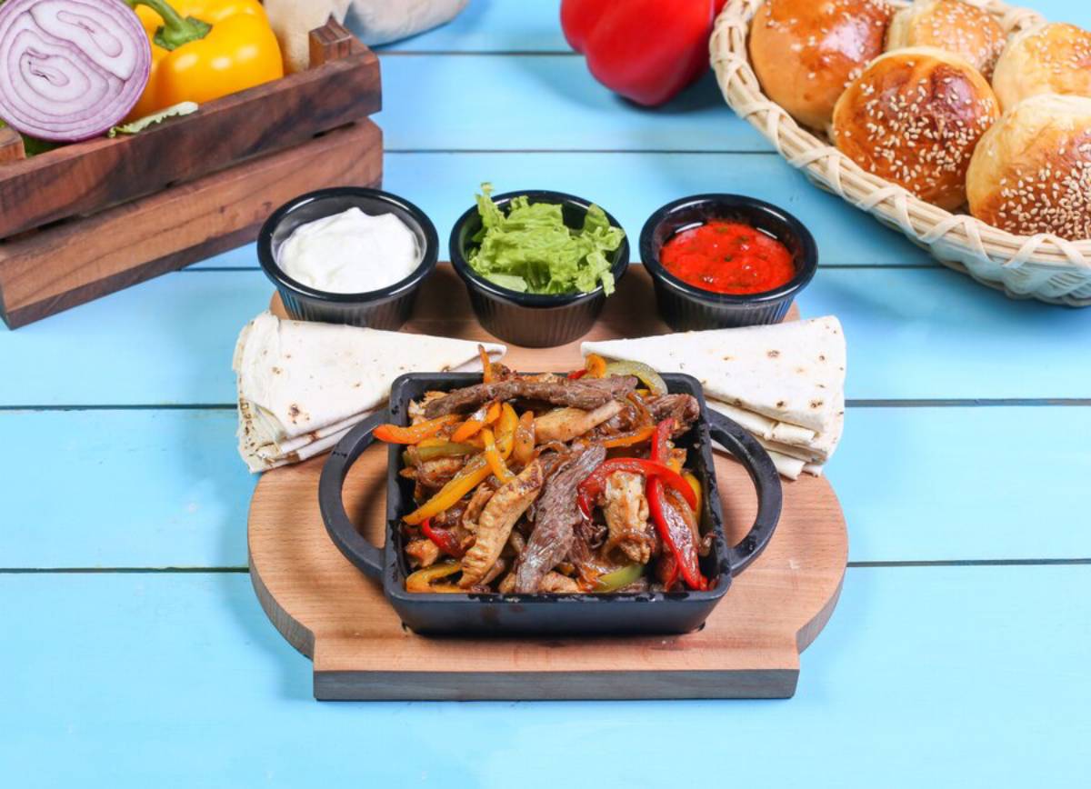 Sizzling beef fajitas with colorful peppers, served with tortillas, lettuce, sour cream, and salsa on a wooden platter.