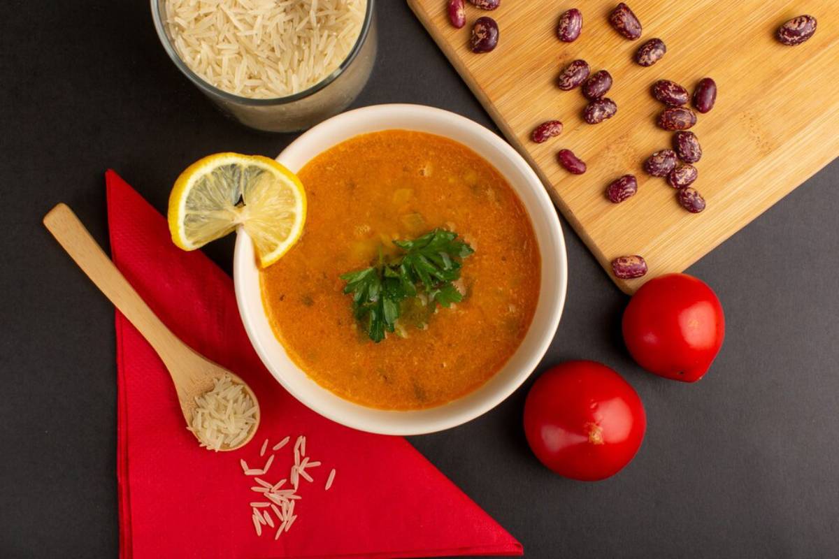 Bowl of rich, orange soup topped with parsley and lemon, accompanied by rice, tomatoes, and kidney beans on a dark background.