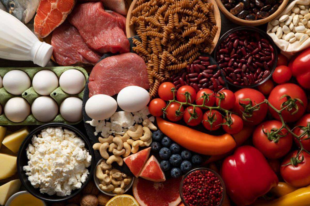 A colorful assortment of fresh foods, including meats, vegetables, fruits, pasta, eggs, nuts, and dairy products arranged artistically.