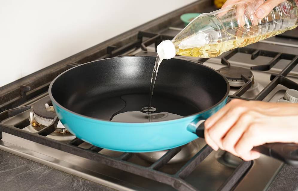 A hand pours oil from a clear bottle into a turquoise frying pan on a stovetop, preparing for cooking.
