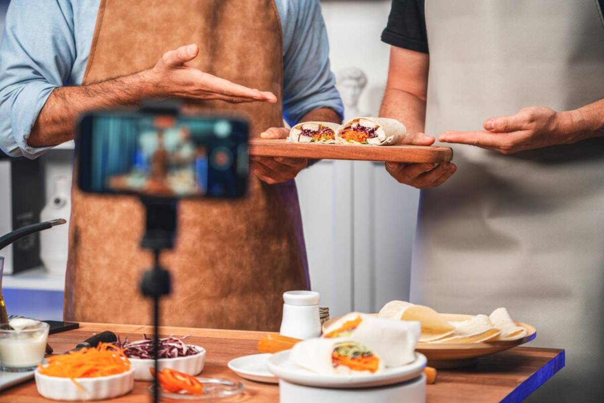 Two chefs in aprons showcase a colorful wrap on a wooden platter while filming with a smartphone, surrounded by fresh ingredients.