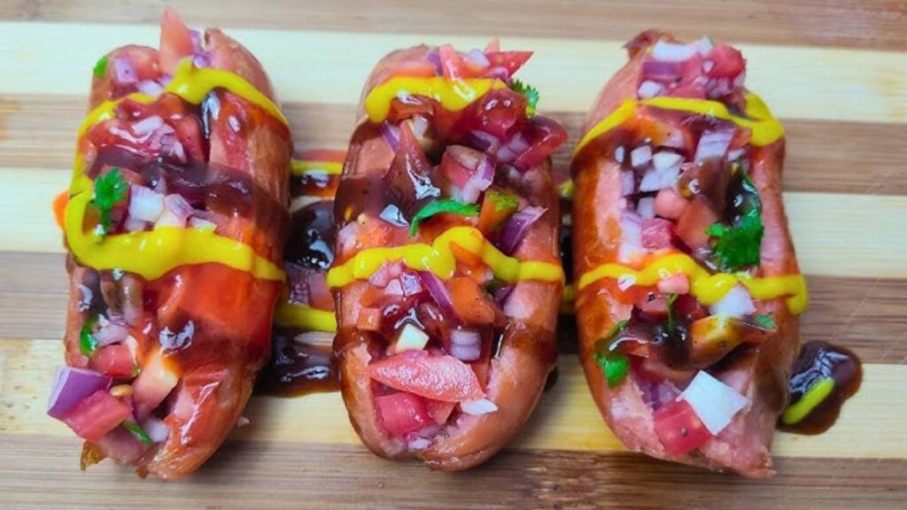 Three vibrant hot dogs topped with diced tomatoes, onions, cilantro, mustard, and barbecue sauce on a wooden surface.
