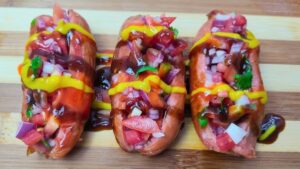 Three vibrant hot dogs topped with diced tomatoes, onions, cilantro, mustard, and barbecue sauce on a wooden surface.
