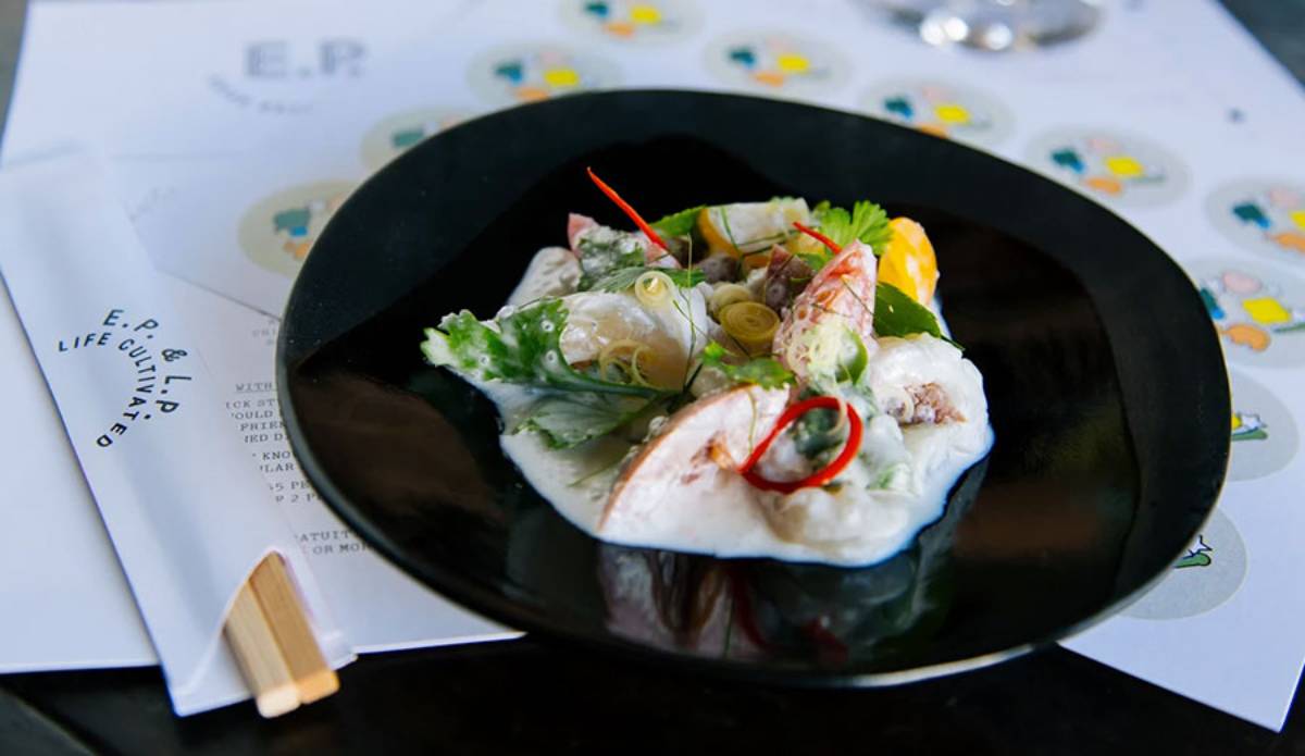 A black plate holds a vibrant seafood dish garnished with herbs, spices, and citrus, alongside a printed menu on the table.