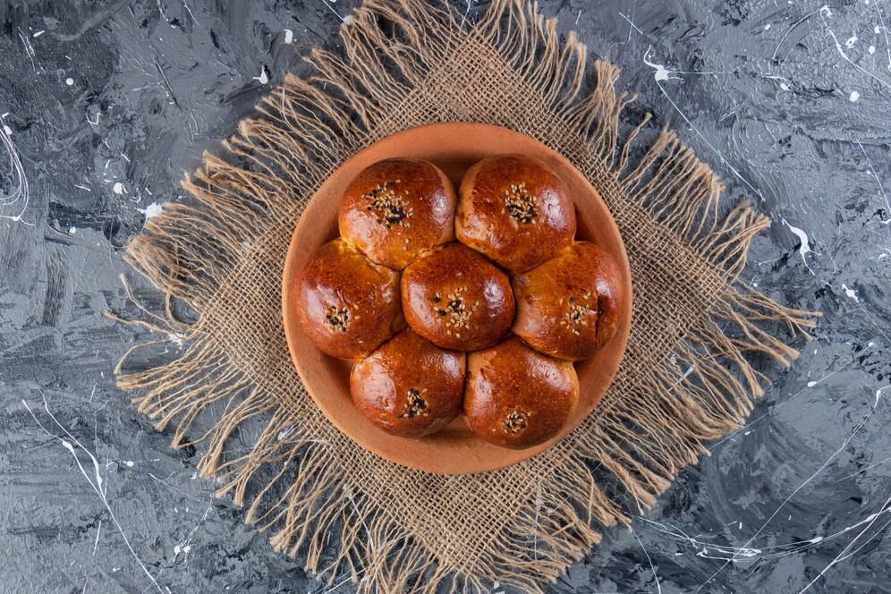 A wooden bowl filled with golden-brown buns topped with sesame seeds, resting on a textured burlap fabric against a dark background.