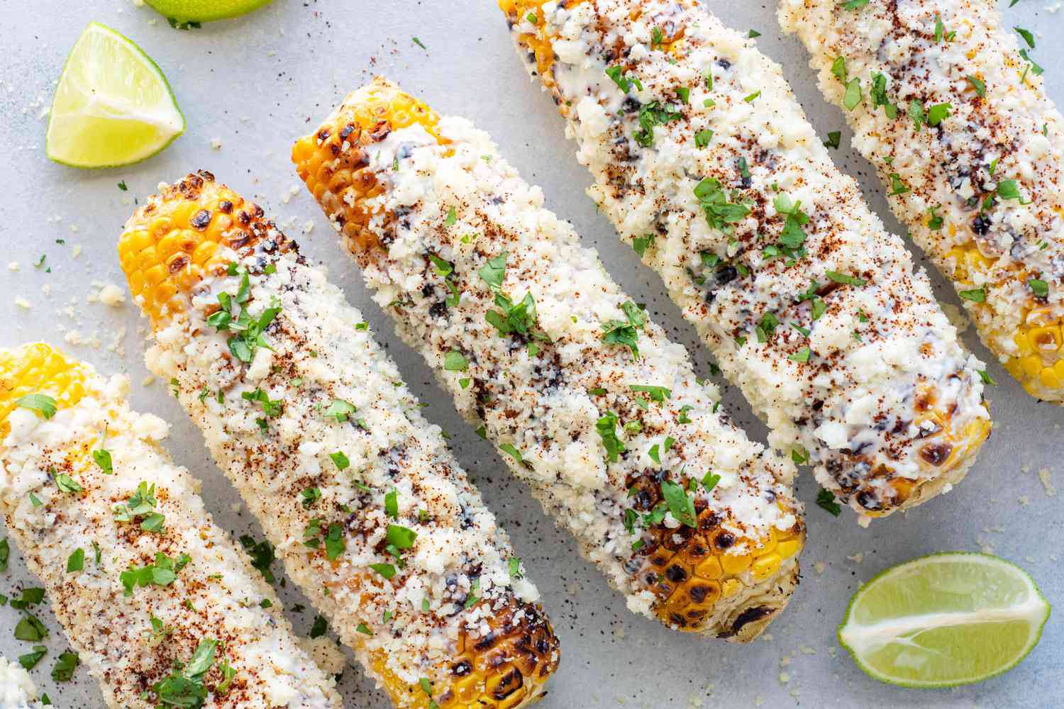 Grilled corn on the cob topped with cheese, spices, and herbs, garnished with lime wedges.