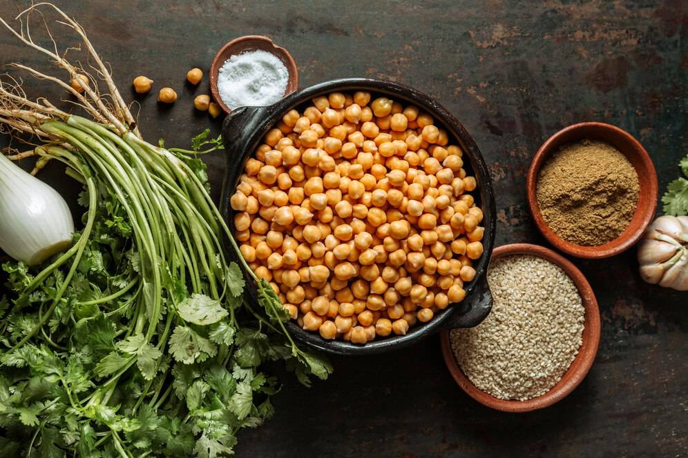 A bowl of chickpeas surrounded by fresh herbs, garlic, and spices on a rustic wooden surface.