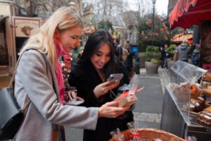 Two women browse items at a festive outdoor market, one holding a coffee cup and the other using a smartphone near a display of treats.