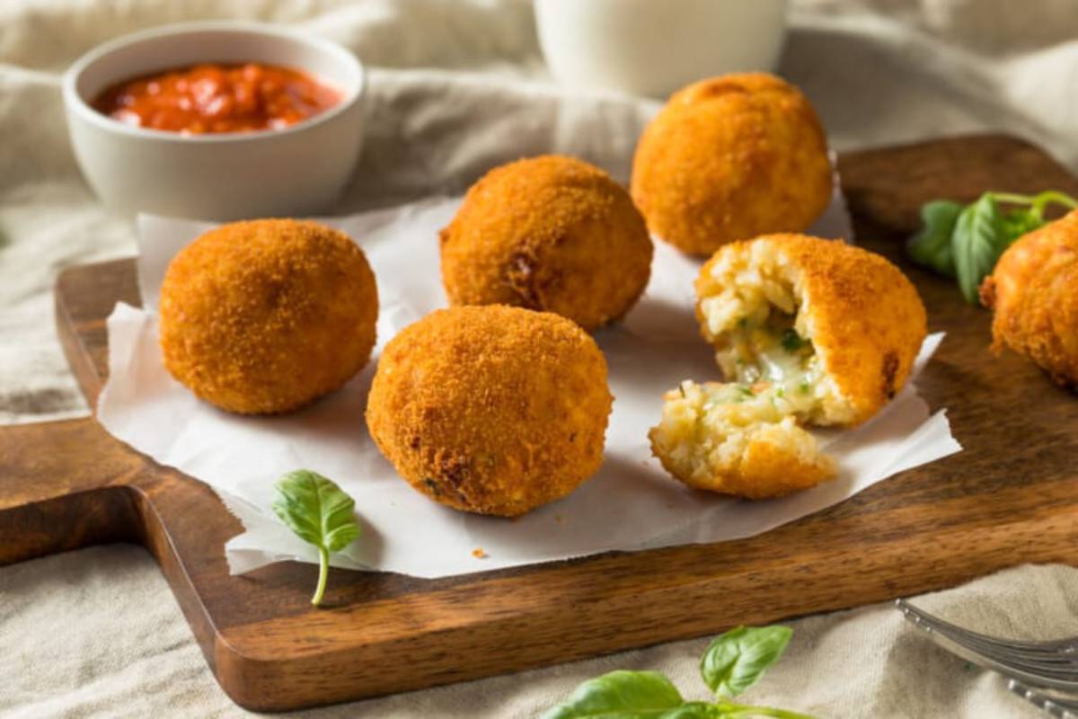 Golden, crispy croquettes on a wooden board, with one partially open to reveal a cheesy, herb-filled interior, accompanied by sauce.