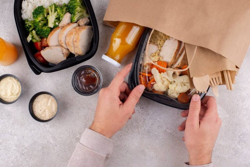 A person is preparing a meal from a takeout box with rice, chicken, vegetables, sauces, and a drink, on a gray countertop.