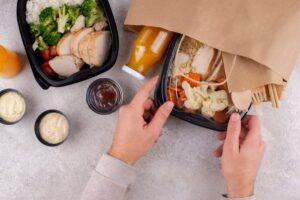 A person is preparing a meal from a takeout box with rice, chicken, vegetables, sauces, and a drink, on a gray countertop.