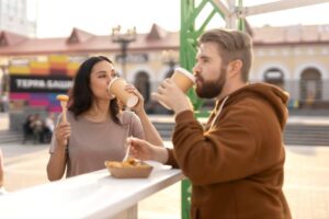 A woman and a man stand at a food stall, enjoying drinks and sharing a plate of fries in a lively outdoor setting.