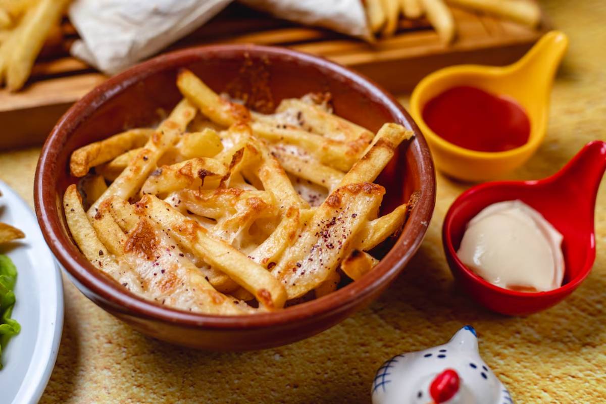 A bowl of golden, crispy fries topped with melted cheese, accompanied by small bowls of ketchup and creamy sauce.