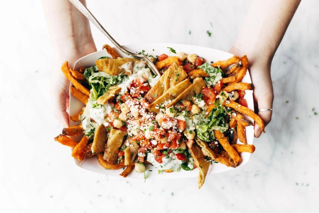 A person holds a bowl of loaded sweet potato fries topped with lettuce, tomato, falafel, chickpeas, and creamy sauce.
