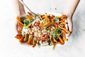 A person holds a bowl of loaded sweet potato fries topped with lettuce, tomato, falafel, chickpeas, and creamy sauce.
