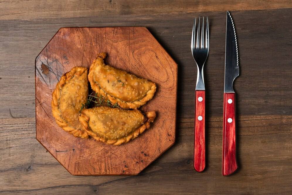 Three golden-brown empanadas on a wooden plate, accompanied by a red-handled fork and knife, on a rustic wooden table.