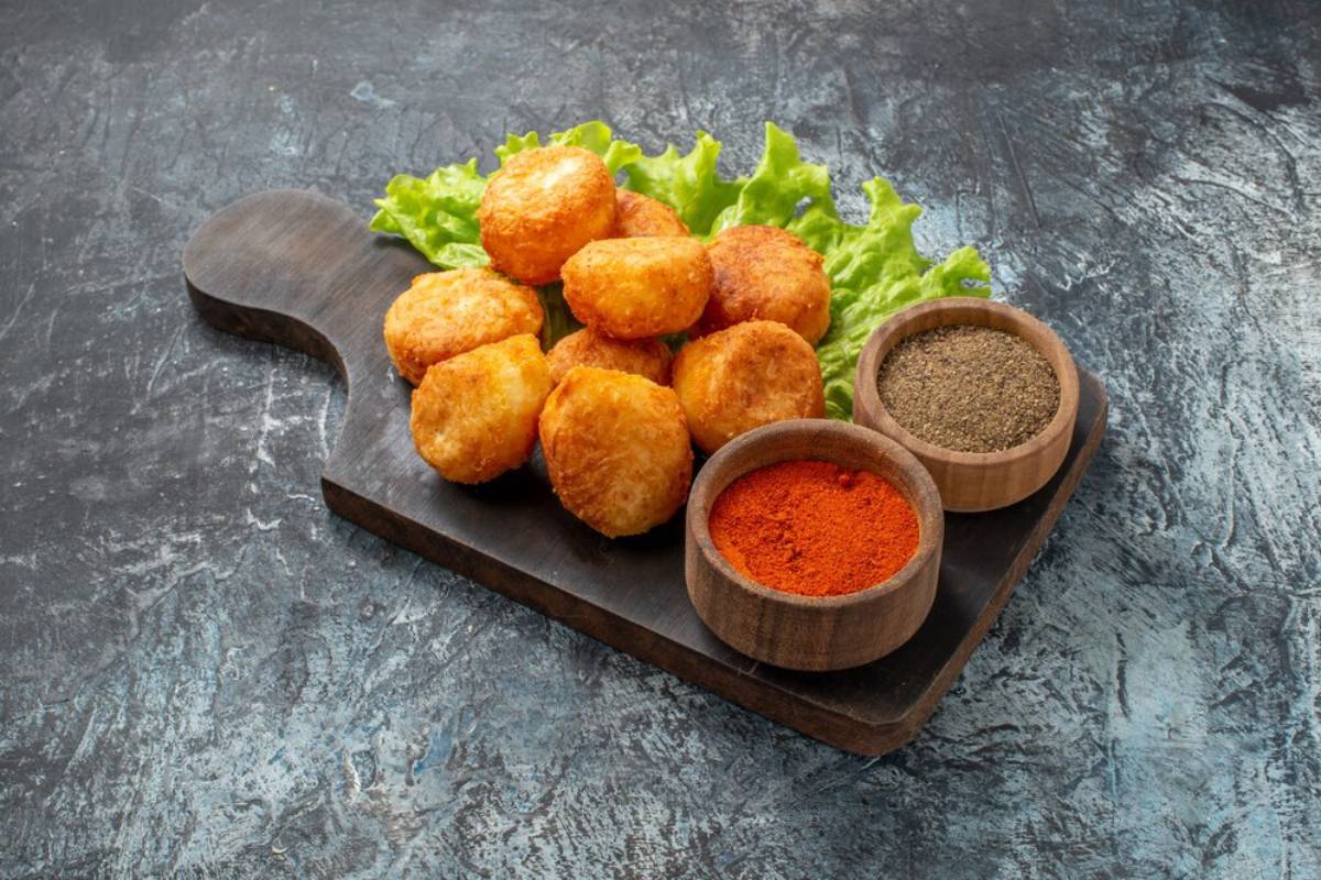 Golden-brown, crispy fried snacks arranged on a wooden board with fresh lettuce and two bowls of spices: red and black pepper.