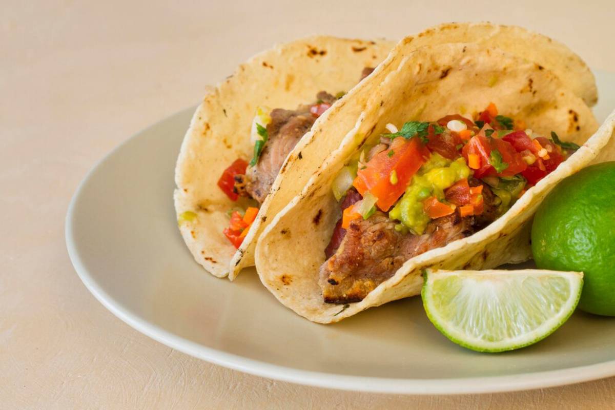 Two soft tacos filled with meat, fresh salsa, and guacamole, served on a plate with a lime wedge.