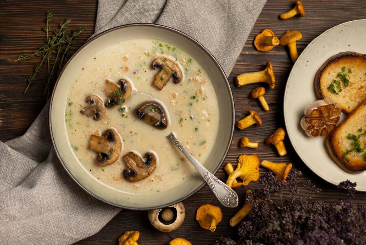 Creamy mushroom soup topped with sliced mushrooms, surrounded by garlic, toasted bread, and fresh herbs on a rustic wooden table.