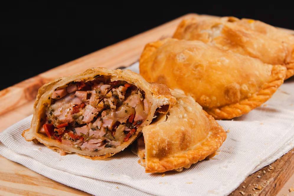 Golden-brown empanadas filled with savory meat, peppers, and herbs, displayed on a wooden board with a napkin underneath.