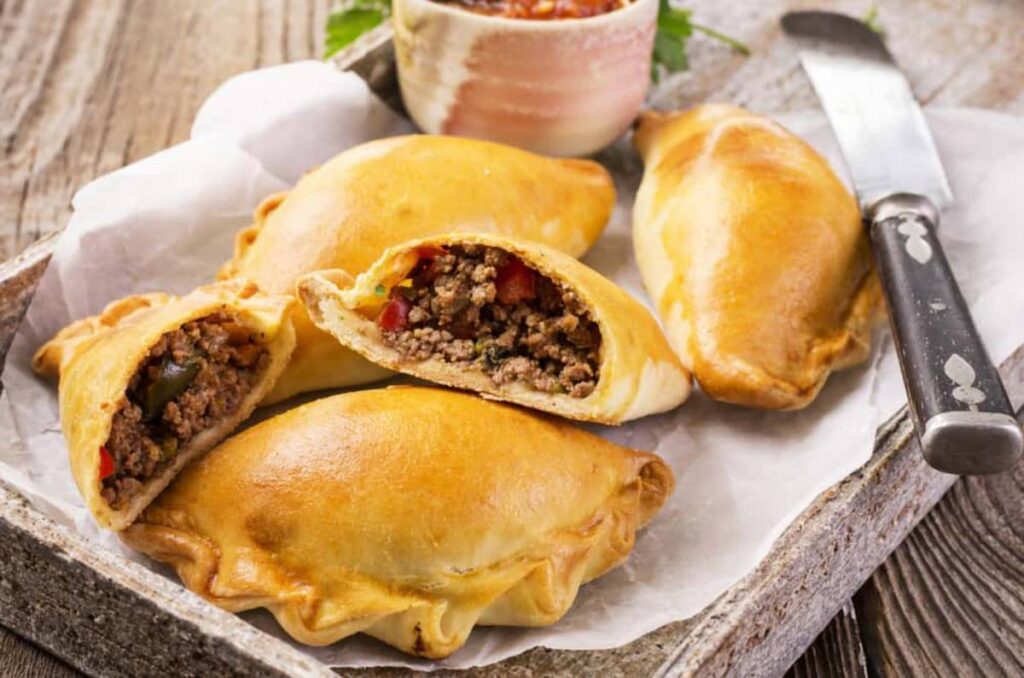 A platter of golden-brown empanadas filled with seasoned ground meat and vegetables, accompanied by a small bowl of salsa.