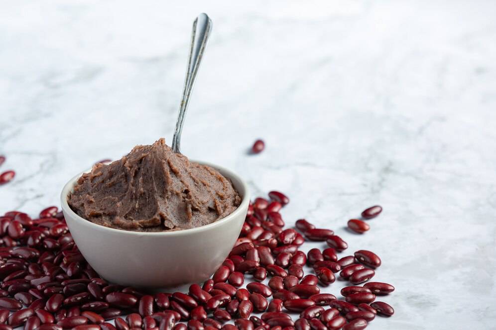 A small bowl of smooth, brown bean paste sits atop a marble surface surrounded by scattered red kidney beans.