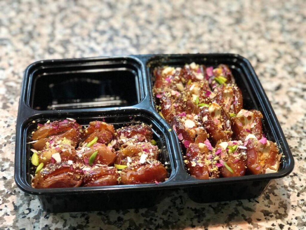 A black container holds two sections filled with stuffed dates, garnished with nuts and colorful flower petals on a textured surface.