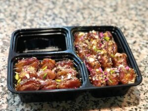 A black container holds two sections filled with stuffed dates, garnished with nuts and colorful flower petals on a textured surface.