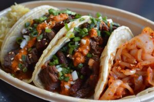 Three loaded tacos with beef, green onions, and spicy sauce, served with a side of kimchi and a scoop of rice.