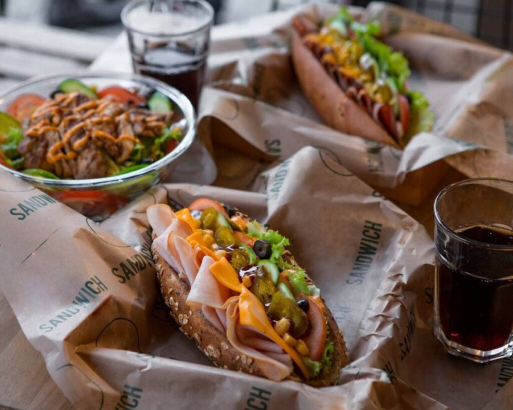 A hearty meal featuring two loaded sandwiches with fresh vegetables, a grilled chicken salad, and glasses of dark beverage on a table.