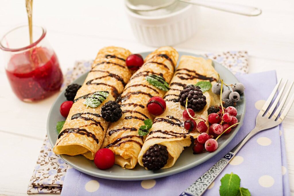 Plated crepes drizzled with chocolate, garnished with mint leaves and fresh berries, alongside a jar of raspberry jam.