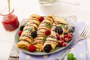 Plated crepes drizzled with chocolate, garnished with mint leaves and fresh berries, alongside a jar of raspberry jam.