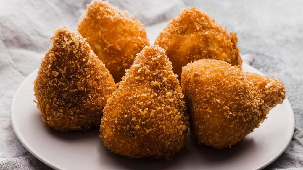 A plate of four golden-brown, triangular croquettes, crispy on the outside and covered in breadcrumbs, resting on a soft fabric.