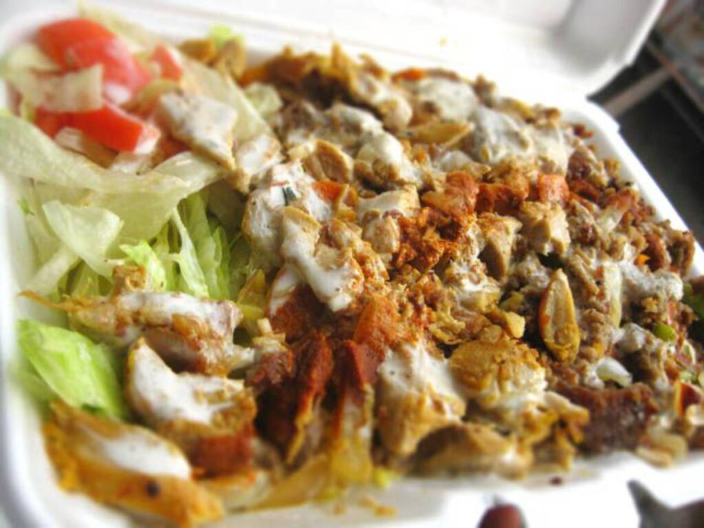 A close-up of a takeout container filled with seasoned grilled chicken, lettuce, tomatoes, and a drizzle of sauce.