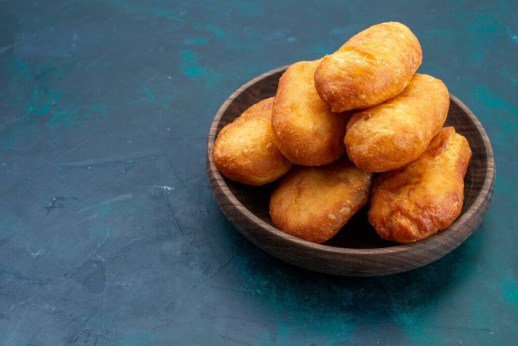 A wooden bowl filled with golden-brown, freshly baked bread rolls stacked on a textured teal surface.