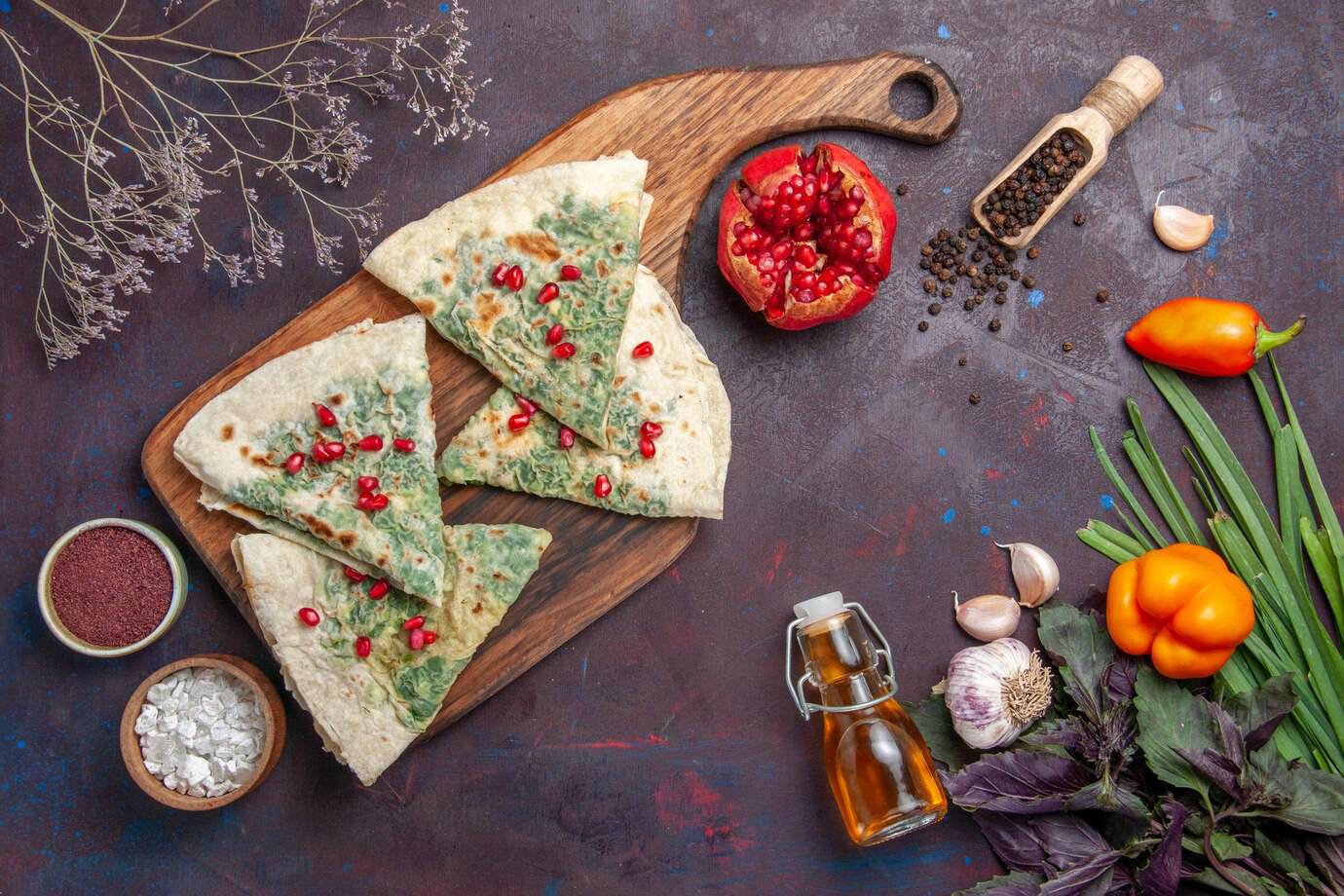 A wooden board holds cut green herb flatbreads topped with pomegranate seeds, surrounded by spices, peppers, garlic, and herbs.
