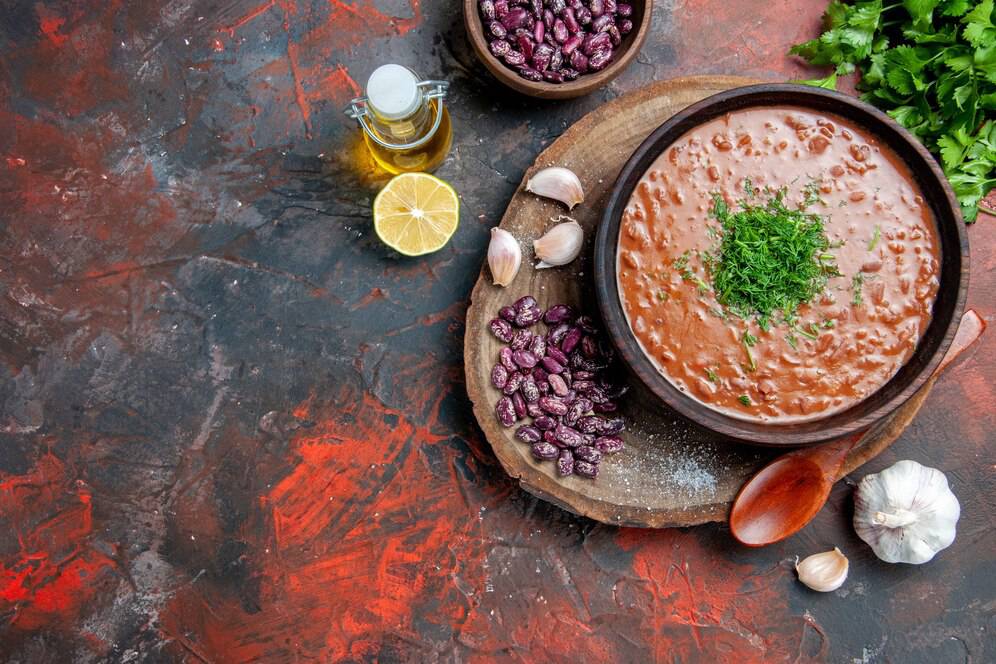 A bowl of hearty bean soup topped with fresh herbs, surrounded by garlic, lemon, olive oil, and purple beans on a rustic table.