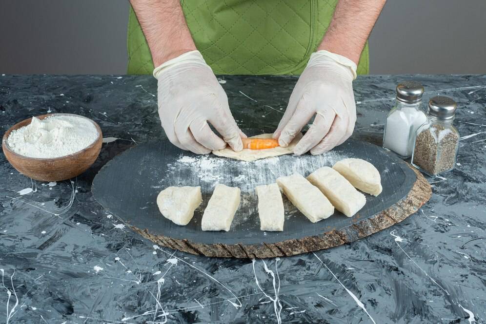 A person in gloves skillfully rolls dough around a piece of carrot on a slate board, with flour and seasonings nearby.