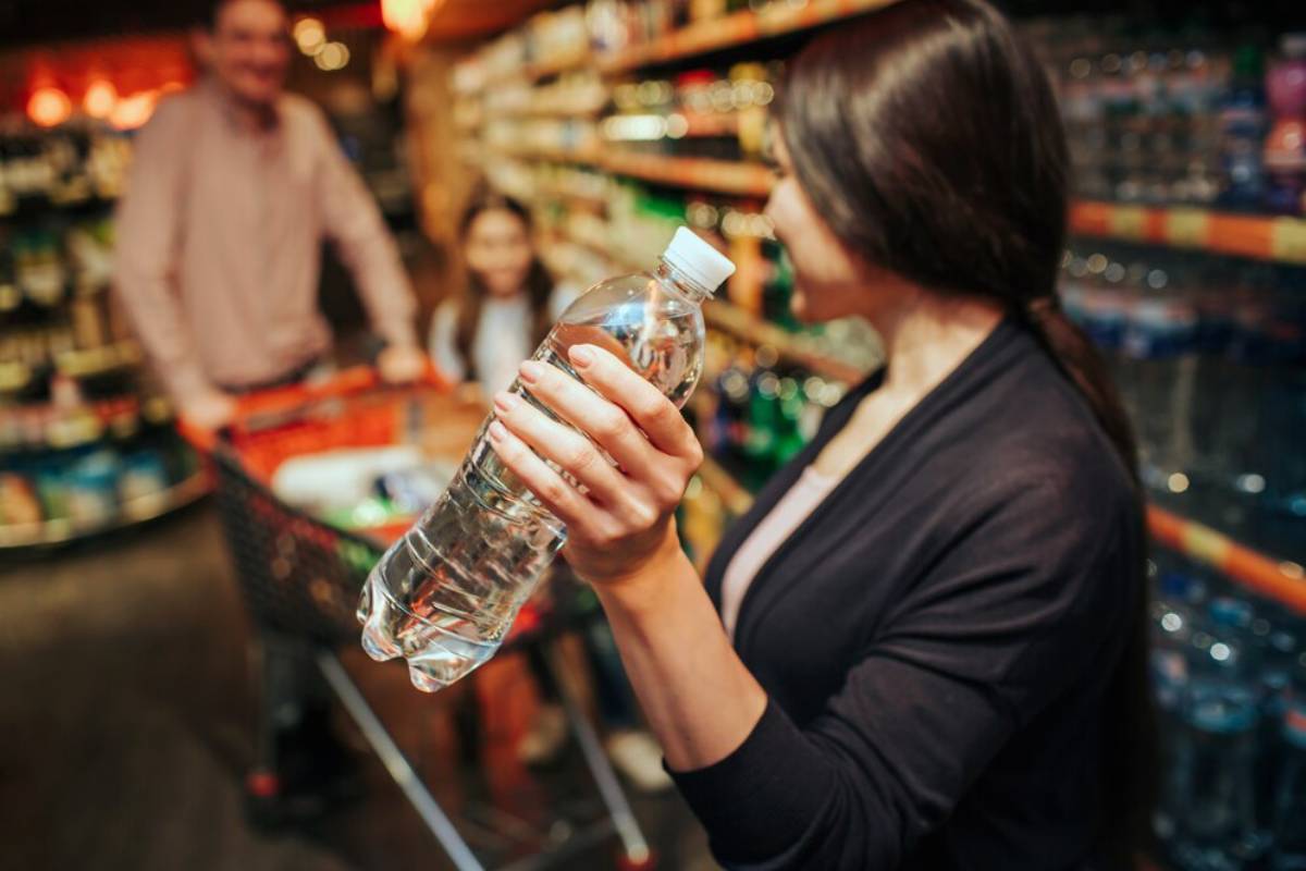 A person holds a clear plastic water bottle in a grocery store aisle, surrounded by colorful beverage displays.